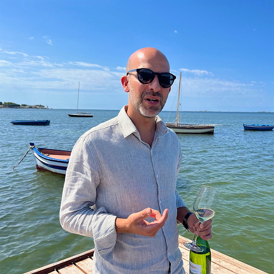 Holding a wine bottle, Pietro Russo stands on the end of a wooden dock.